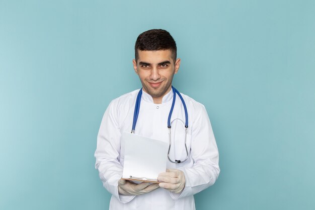 Young male doctor in white suit with blue stethoscope holding notepad and smiling