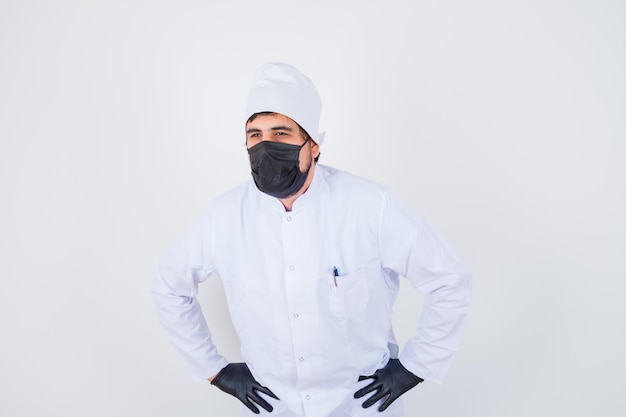 Young male doctor in white uniform holding hands on waist and looking wistful , front view.