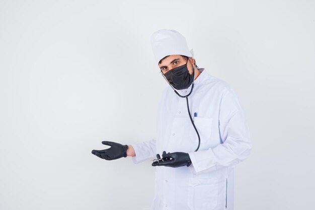 Young male doctor in white uniform pretending to show something and looking confident , front view.