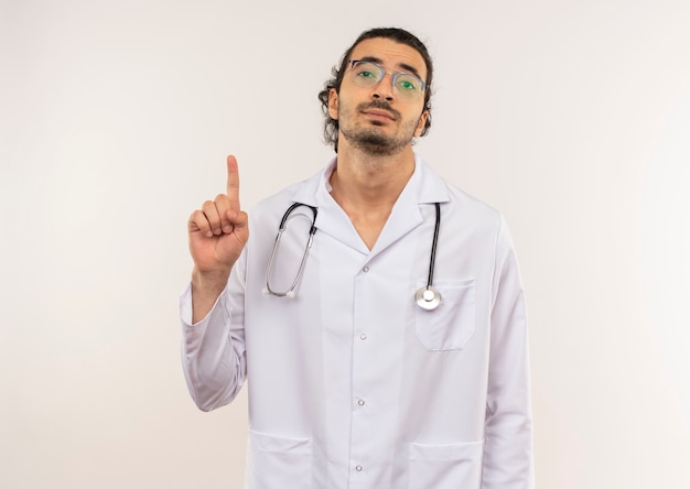 Молодой мужчина-врач в оптических очках в белом халате со стетоскопом указывает вверх