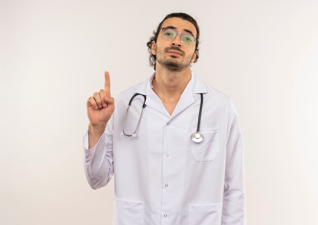 young male doctor with optical glasses wearing white robe with stethoscope points to up