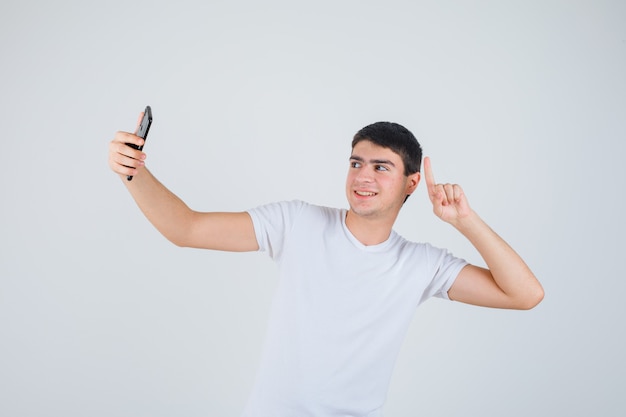 Young male doing selfie while pointing up in t-shirt and looking cheery , front view.