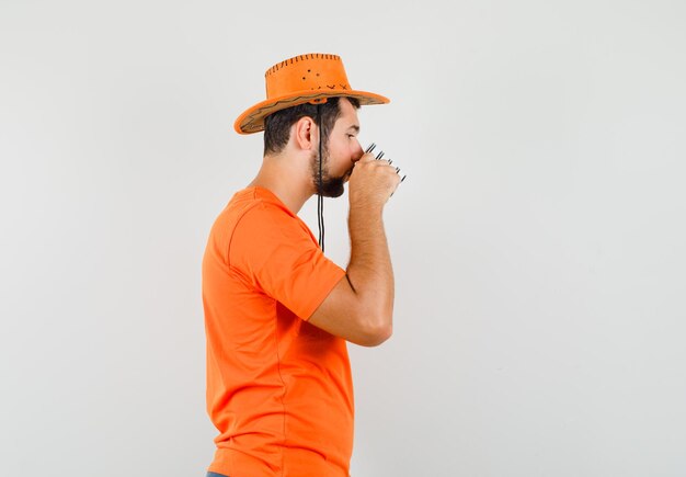 Бесплатное фото Молодой мужчина пьет чай в оранжевой футболке, шляпе и выглядит мило. .