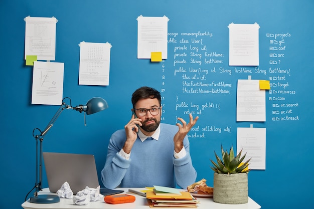 Free photo young male entrepreneur thinks on business solution during telephone conversation, raises hand confusingly, sits at white desk with notepads, crumpled paper and laptop computer