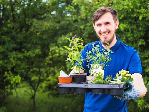 Young male gardener holding crate with vivid potted plants in the garden