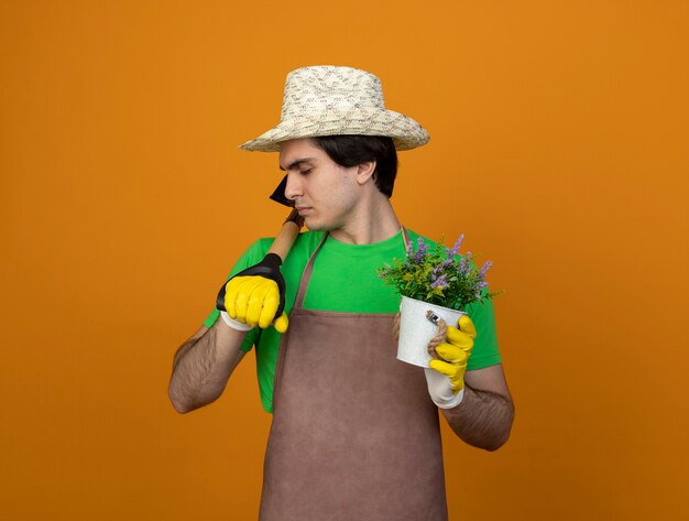 Young male gardener in uniform wearing gardening hat with gloves with closed eyes holding flower in flowerpot putting spade on shoulder isolated on orange