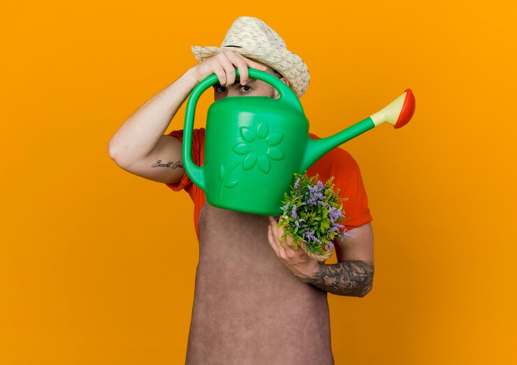 Free Photo young male gardener wearing gardening hat holds flowers in flowerpot and looks through watering can 