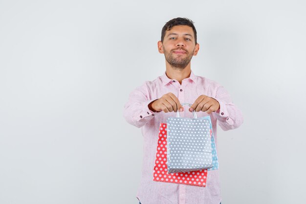 Young male giving you paper bags in shirt and looking cheerful , front view.