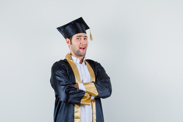 Young male in graduate uniform standing with crossed arms and looking proud , front view.