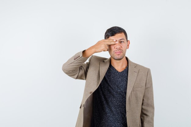 Young male in grayish brown jacket,black shirt closing his one eye with hand and looking weird , front view.