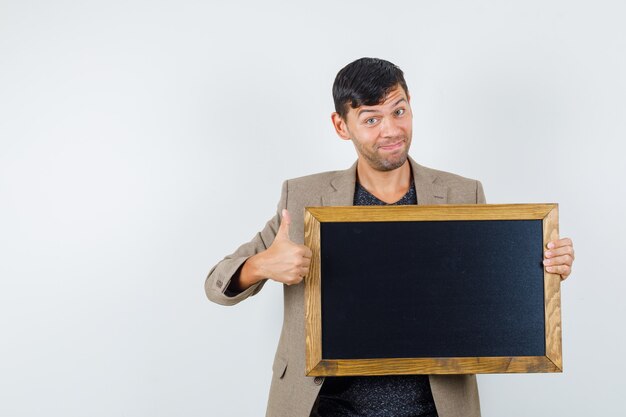 Young male in grayish brown jacket,black shirt holding cardboard while showing thumb up and looking pleased , front view.