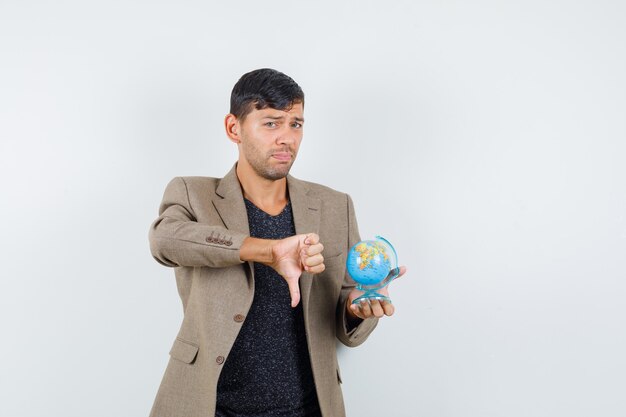 Young male in grayish brown jacket,black shirt holding mini globe while showing thumb down and looking displeased , front view.