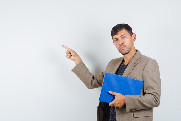 Young male in grayish brown jacket,black shirt holding notebook while pointing aside and looking confident , front view.