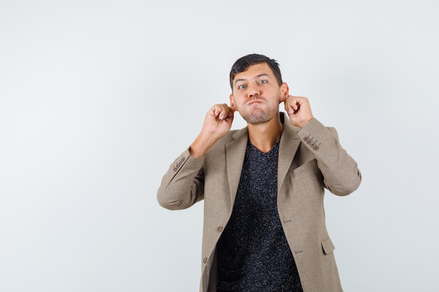 Young male in grayish brown jacket,black shirt pulling his ears while puffing cheeks and looking weird , front view.