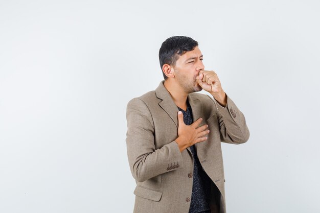 Young male in grayish brown jacket coughing and looking uncomfortable , front view.