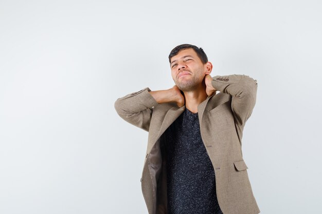 Young male in grayish brown jacket holding hand on neck and looking tired , front view.