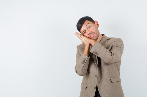 Young male in grayish brown jacket standing while making pillow gesture and looking awake , front view.