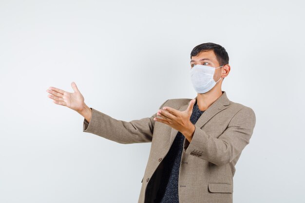 Young male in grayish brown jacket,white mask showing something and looking amazed , front view.