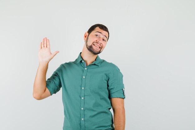 Young male in green shirt waving hand for greeting and looking joyful , front view.