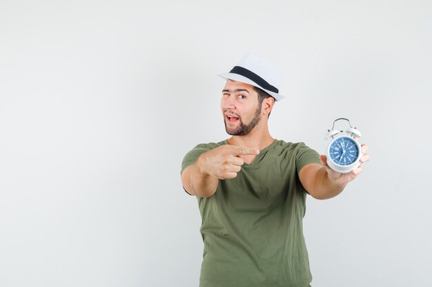 Young male in green t-shirt and hat pointing at alarm clock and winking eye