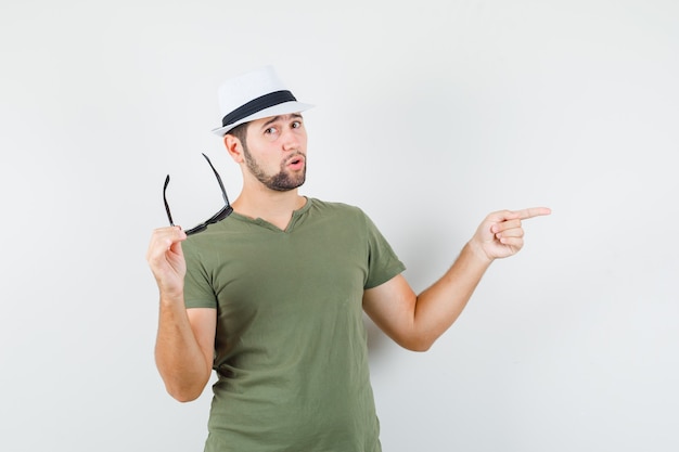 Young male in green t-shirt and hat pointing away while holding glasses and looking cool