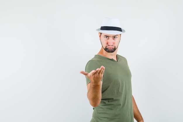 Young male in green t-shirt and hat raising hand in questioning manner and looking weird