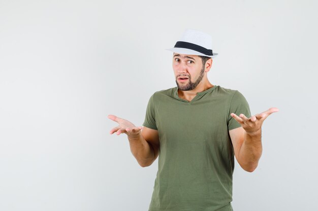 Young male in green t-shirt and hat raising hands in questioning manner and looking confused