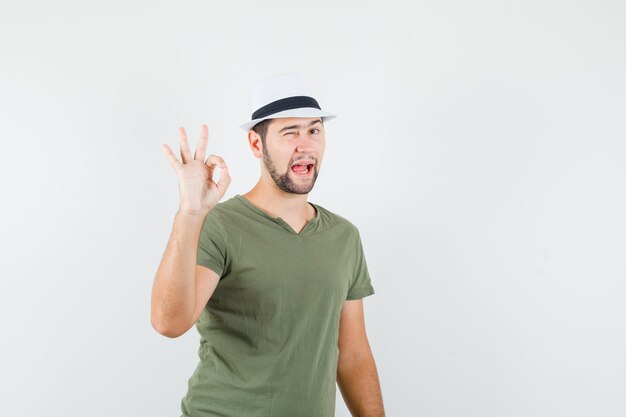 Молодой мужчина в зеленой футболке и шляпе показывает знак ОК и подмигивает