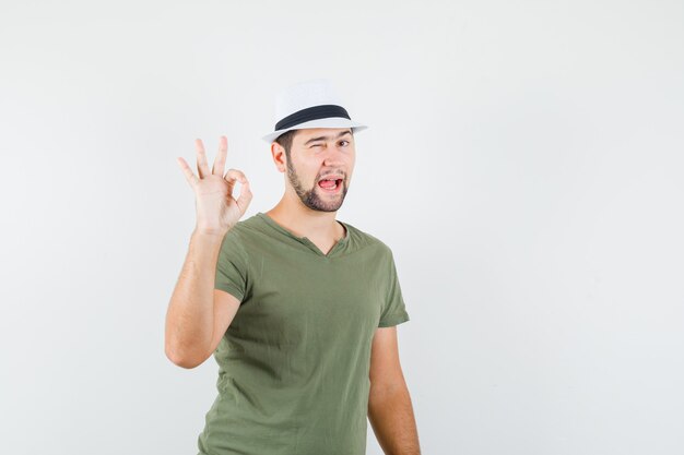 Young male in green t-shirt and hat showing ok sign and winking eye