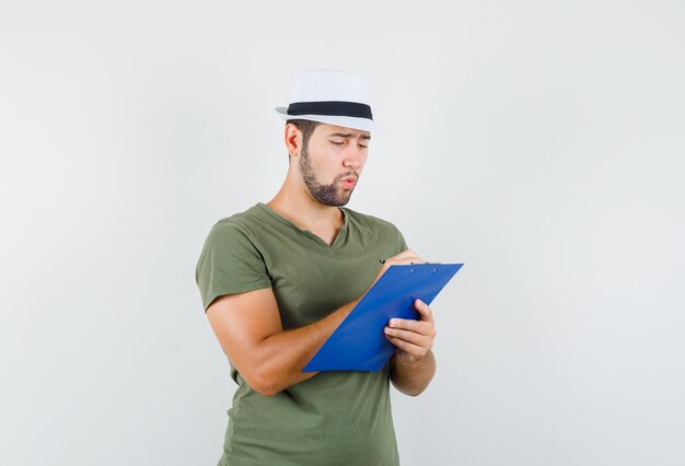 Young male in green t-shirt and hat taking notes on clipboard and looking busy
