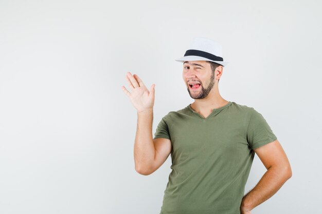 Young male in green t-shirt and hat waving hand, winking eye, sticking out tongue and looking funny