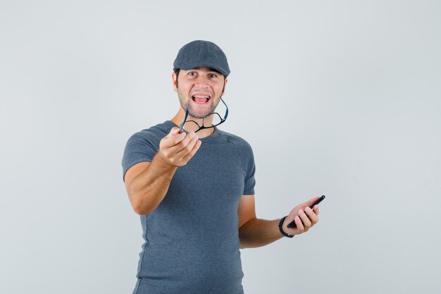 Young male in grey t-shirt cap holding mobile phone and glasses and looking jolly  