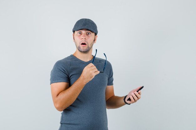 Молодой мужчина в серой кепке футболки держит мобильный телефон и очки и выглядит удивленным