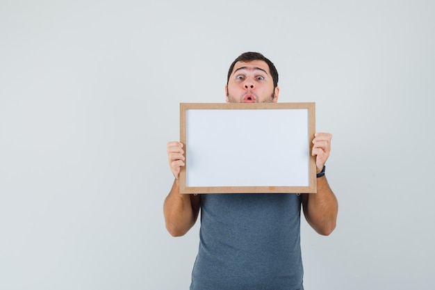 Young male in grey t-shirt holding empty frame and looking surprised  