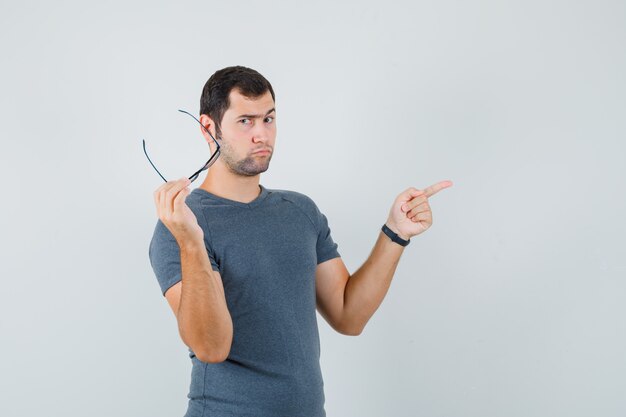 Young male in grey t-shirt holding glasses pointing aside and looking pensive  