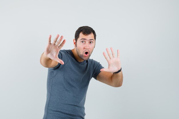 Young male in grey t-shirt holding hands to defend himself and looking scared  