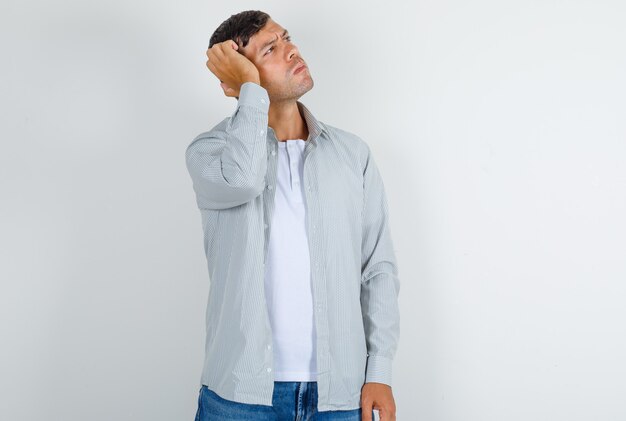 Молодой мужчина в серой футболке, джинсах смотрит вверх с рукой на голове и выглядит задумчивым