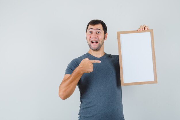 Young male in grey t-shirt pointing at empty frame and looking cheerful  