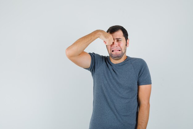 Young male in grey t-shirt rubbing eye while crying and looking offended  