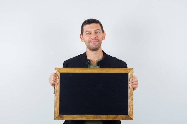 Young male holding blackboard in t-shirt, jacket and looking cheerful. front view.