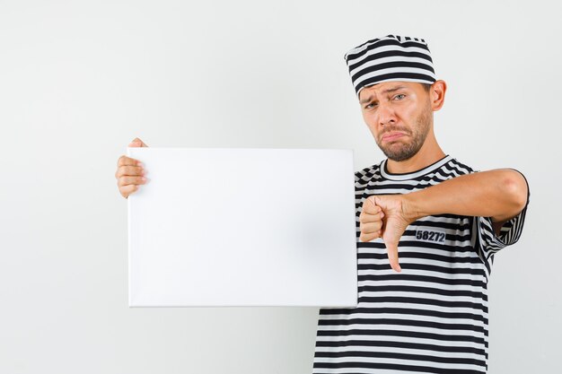 Young male holding blank canvas showing thumb down in striped t-shirt hat and looking sad  