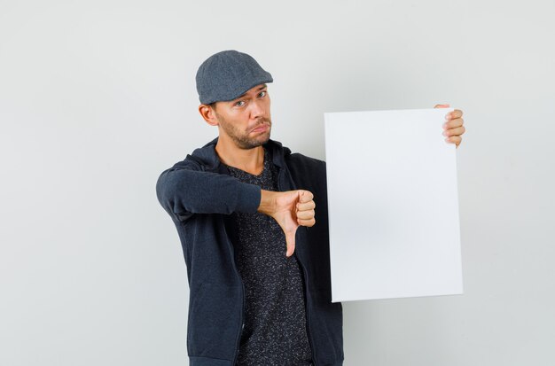 Young male holding blank canvas, showing thumb down in t-shirt, jacket, cap front view.