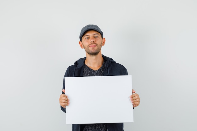 Young male holding blank canvas and smiling in t-shirt, jacket, cap front view.