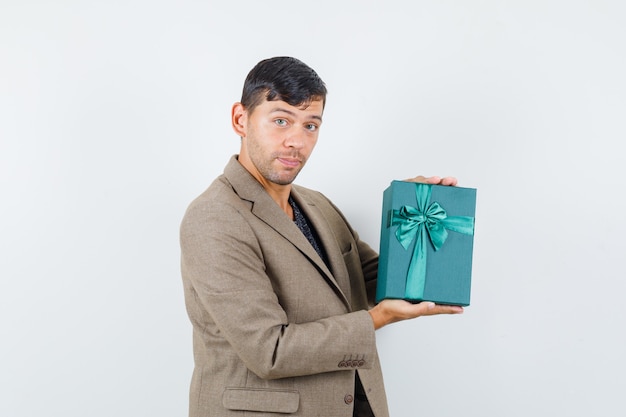 Young male holding blue gift box in grayish brown jacket and looking confident , front view.