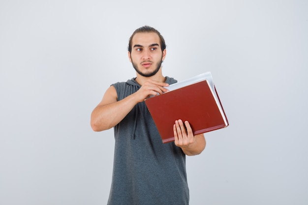 Young male holding book while looking away in sleeveless hoodie and looking pensive. front view.