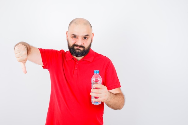 Young male holding bottle while showing thumb down in red shirt and looking dissatisfied. front view.