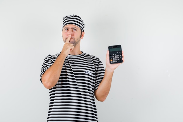 Молодой мужчина держит калькулятор в футболке, шляпе и нерешительно смотрит. передний план.