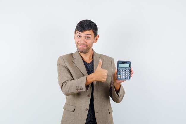 Young male holding calculator while showing thumb up in grayish brown jacket,black shirt and looking glad , front view.