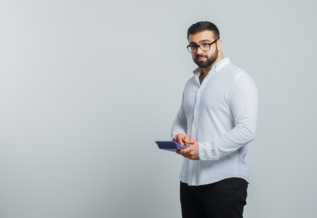 Young male holding calculator in white shirt, pants and looking pensive