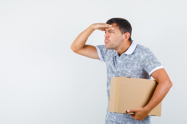 Young male holding cardboard box with hand over eyes in t-shirt and looking watchful